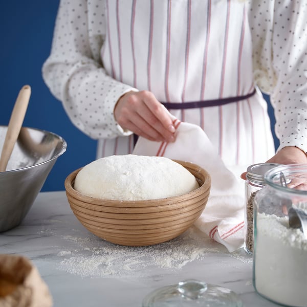 A person is placing dough into a round wooden proofing basket dusted with flour, surrounded by utensils and ingredients.
