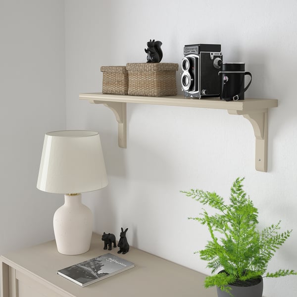 BERGSHULT shelf in beige holds baskets, camera, mug, decor. Light wood, minimalist design above table with lamp and plants.