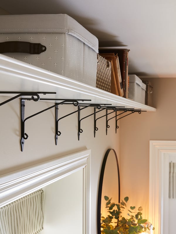 White wall shelf with bevelled edge, dark brackets, storage boxes, plant, mirror.
