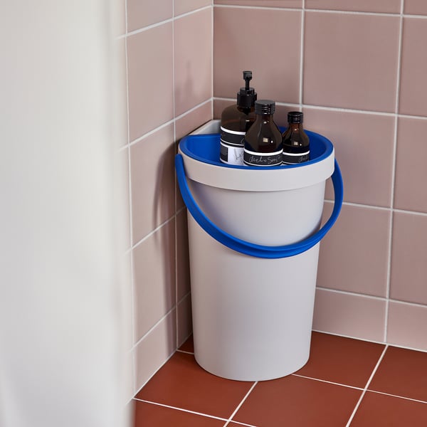 A corner shower bucket with blue handles collecting water in a pale pink tiled bathroom.