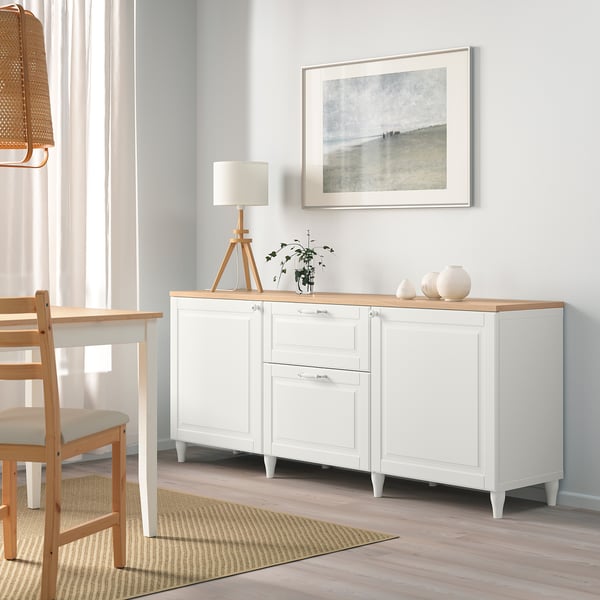 White BESTÅ sideboard with adjustable shelves, drawers, light wooden top, seascape art, wooden chair, curtained window.