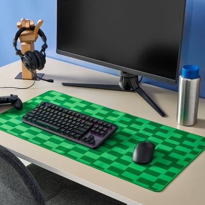 Gaming setup with large green chequered BLÅSKATA mouse pad. Keyboard, mouse, monitor, headphones, and water bottle on a grey desk.