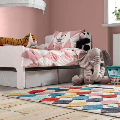 Kids room with BUSENKEL rug, colourful harlequin pattern, soft and durable, fits under bed, surrounded by plush toys, pink walls.