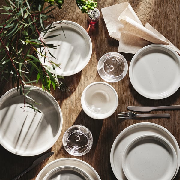 White stoneware plate set on wooden table with glasses, cutlery, and plant. Plates are larger than glasses and napkins.