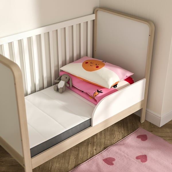 Small, white baby bed with pink and white decorated mattress, stuffed toy, and heart-shaped rug. Modern, minimalist design.