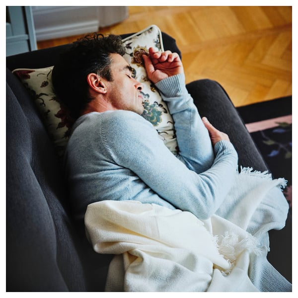 Person relaxing on soft, grey ESSEBODA sofa with floral pillow, draped in white blanket.