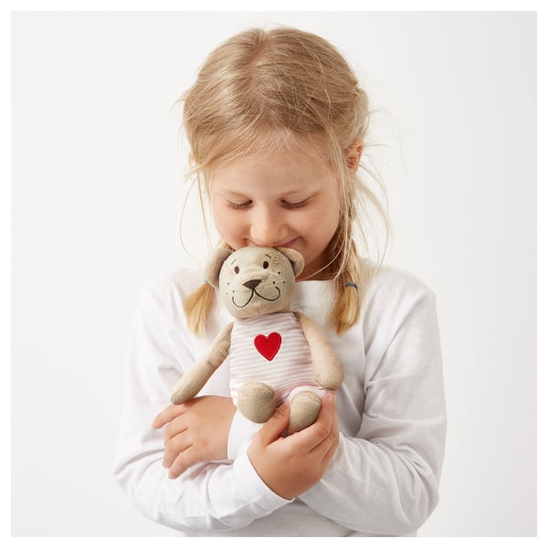 A young girl holding a beige teddy bear with a heart on its white striped shirt.