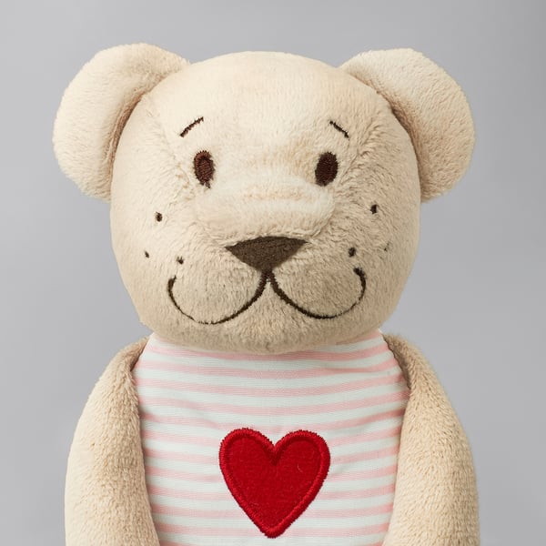 A beige teddy bear with a red heart-shirt, soft and plush, smiling. Ideal for cuddling and play.