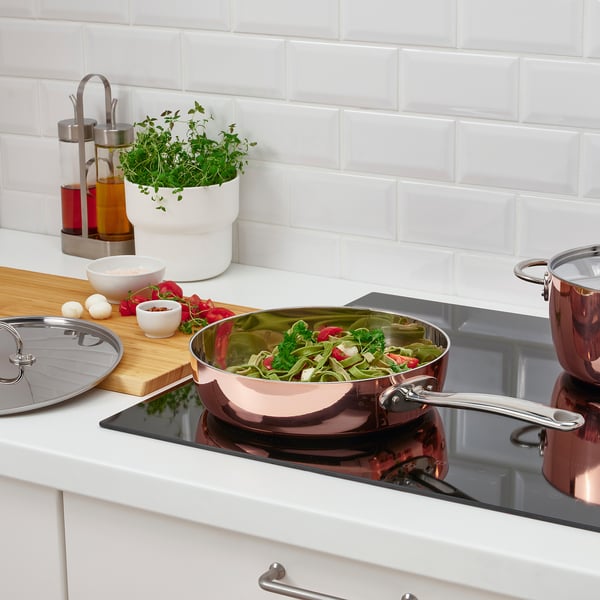Modern kitchen with copper pan on induction hob, vegetables, and broth. Wooden board with ingredients and plant.