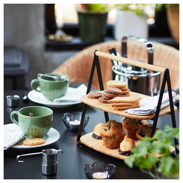 Two-tiered bamboo serving tray holds cookies and muffins next to green tea cups on a dark wooden table.