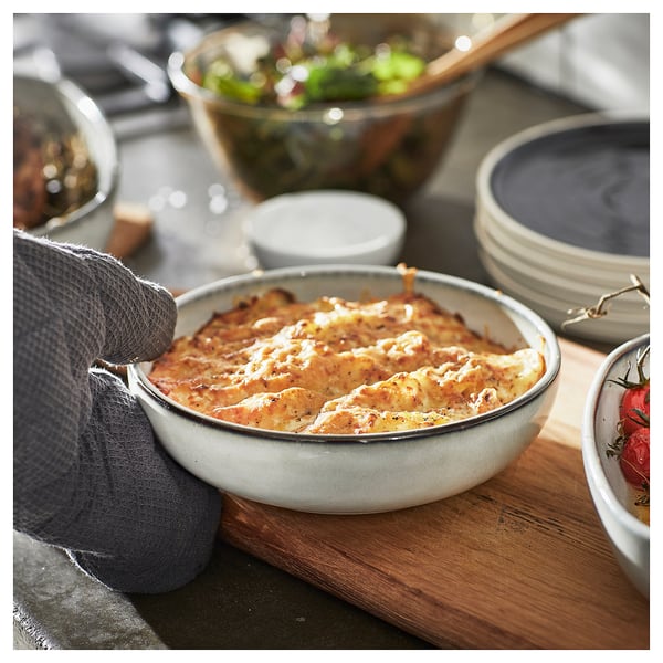 A hand in a grey oven mitt holds a blue ceramic dish with lasagna, surrounded by bowls and a plate with tomatoes and herbs on a wooden board.