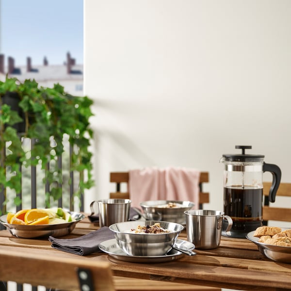 Outdoor table with GRILLTIDER dishes: plates, bowls, mugs. Serving tray has fruit, coffee press, cookies. Medium, stackable.