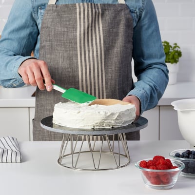 Person icing cake with green spatula. Cake on metal stand, berries nearby.