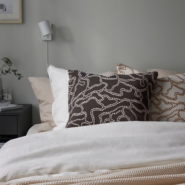 Bed with GULDFLY anthracite cushion, pure cotton, jacquard pattern, wall-mounted lamp, vase, book, light blue wall.
