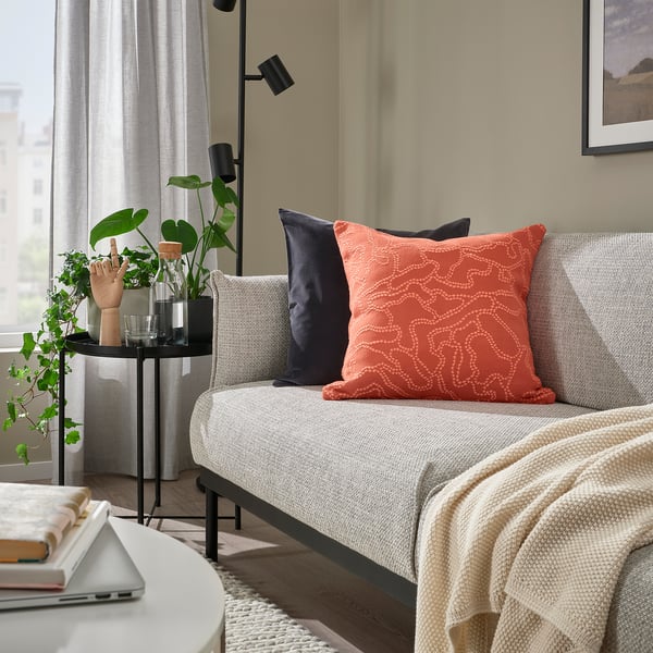 Modern living room with grey sofa, orange GULDFLY cushion, wooden floor, and indoor plant. Cushion is square and patterned.