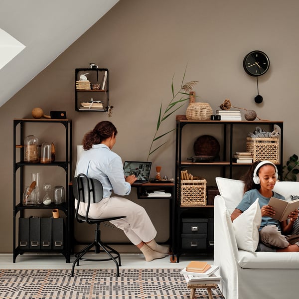 Home office: two people working, one on laptop, one reading with headphones. Cosy decor with plants and clock.