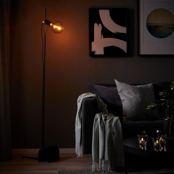 Modern living room with grey HÅRSLINGA floor lamp, sofa, pillows, blankets, candles, and art on dark walls.
