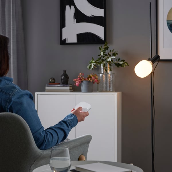 Person sits in grey chair, holding remote aiming at lit floor lamp with round bulb, white cabinet with vases and books behind.
