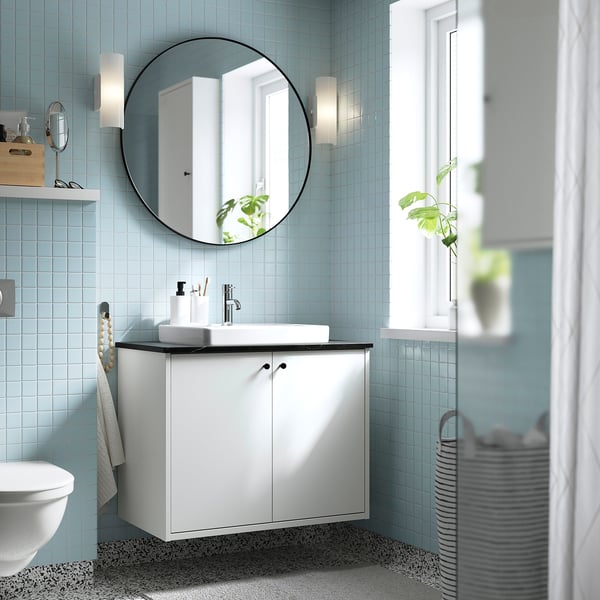 Modern bathroom with HAVBÄCK white cabinet. Round mirror above square sink. Light blue tiles. Plants and towels visible.