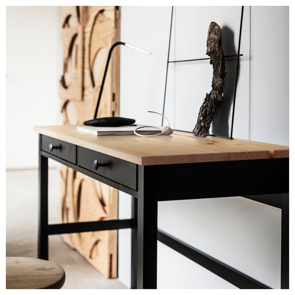 A stylish HEMNES black desk with a light wood top. It has two drawers and a minimalist design, showcasing a lamp, book, and decorative item.