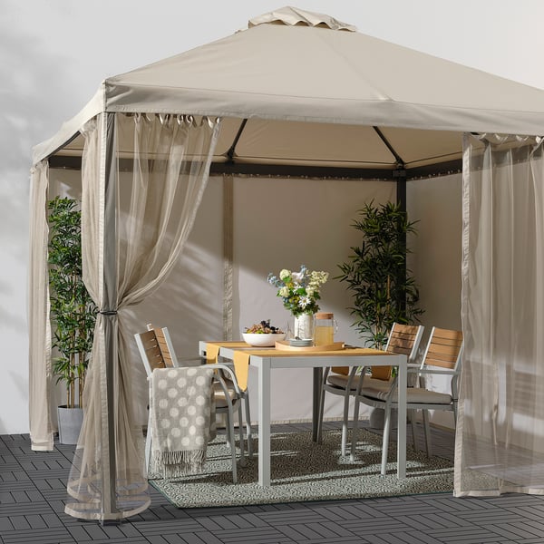 Beige gazebo with sheer polyester curtains, fitting multiple chairs and table, creating cosy ambiance.