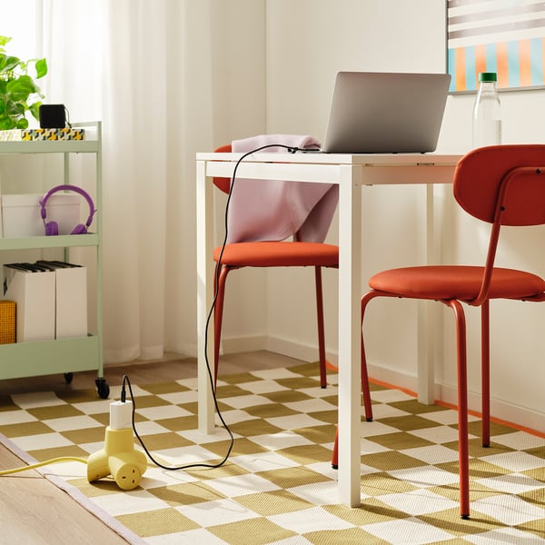 A white desk with two red-orange chairs, a laptop, a water bottle, and a playful yellow 4-way extension cord on the floor providing power to devices.