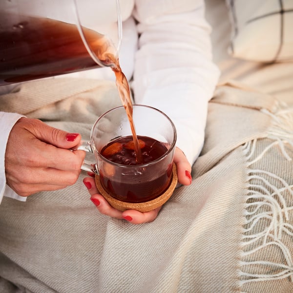 A person pours coffee into a glass mug on a beige cork coaster, protecting the table from heat and reducing noise.