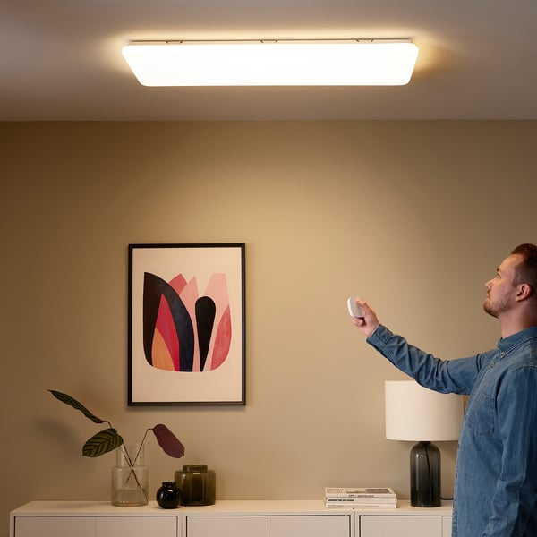 Person uses remote to adjust smart light panel on ceiling, illuminating room. Art, plant, and lamp on sideboard below.