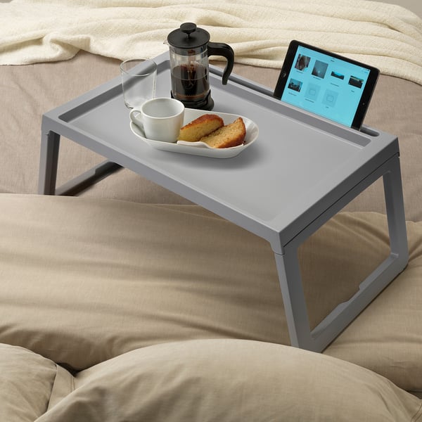 A grey bed tray holds a coffee maker, cup, food, and tablet on beige bed linens.