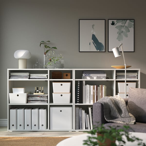 Modern living room with KUGGIS boxes on shelf, books, plants. Art above, sofa, and lamp.