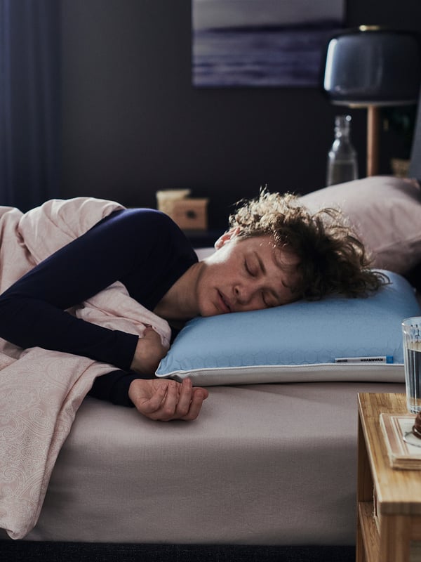A person sleeps on a bed with a blue KVARNVEN pillow. Next to the person is a glass on a wooden bedside table. Beside the person, theres a lamp.