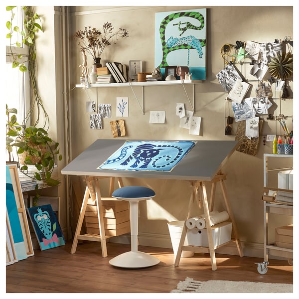 Art studio with LAGKAPTEN desk, light wood, blue stool, shelves with art supplies, plants, posters, and abstract paintings.