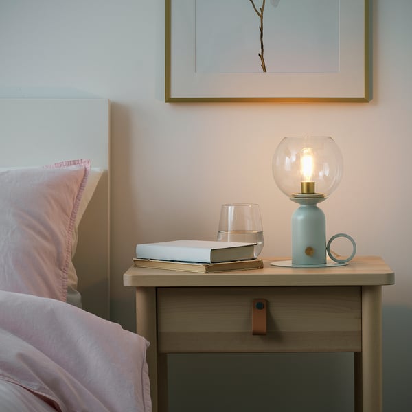 Bedside bedside table with light blue lamp, clear bulb & warm light, partially lit book, wine glass, near headboard.