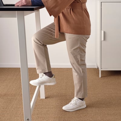 A person leaning on a high table with one foot on a built-in footrest, demonstrating its use for both sitting and standing work.