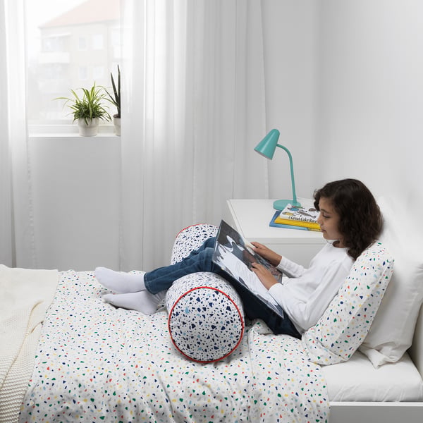 Child reading on colourful bed, lit by lamp, with plant. Wearing white shirt, blue jeans, socks.