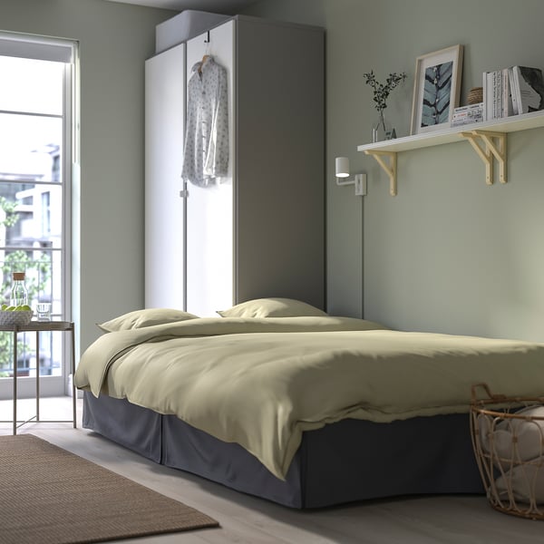 Modern bedroom with bed that converts to sofa, beige bedding, grey wall, bookshelf.
