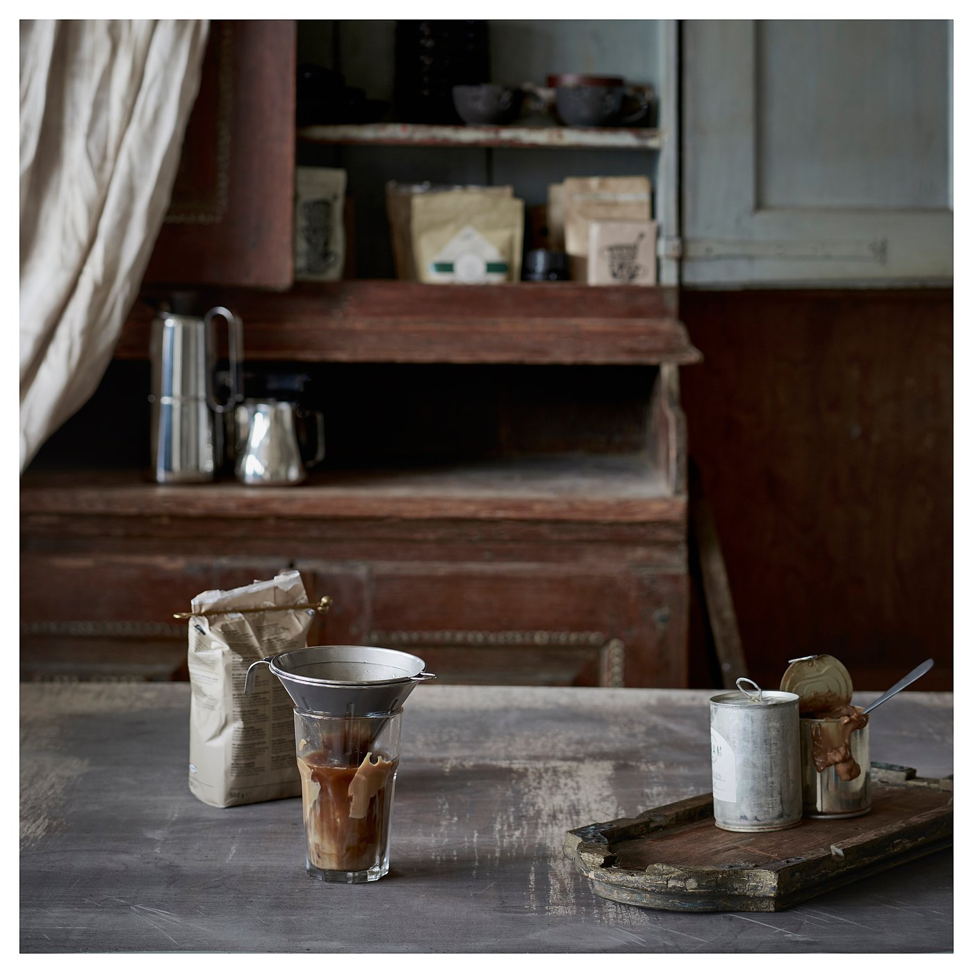 Vintage kitchen with coffee setup, including a silver coffee philtre, bagged beans, and containers with utensils on a rustic wooden table.