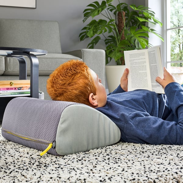 Child lies on floor, reading book, supported by grey ÖVNING footrest and cushion. Beside grey sofa, near potted plant and books on side table.