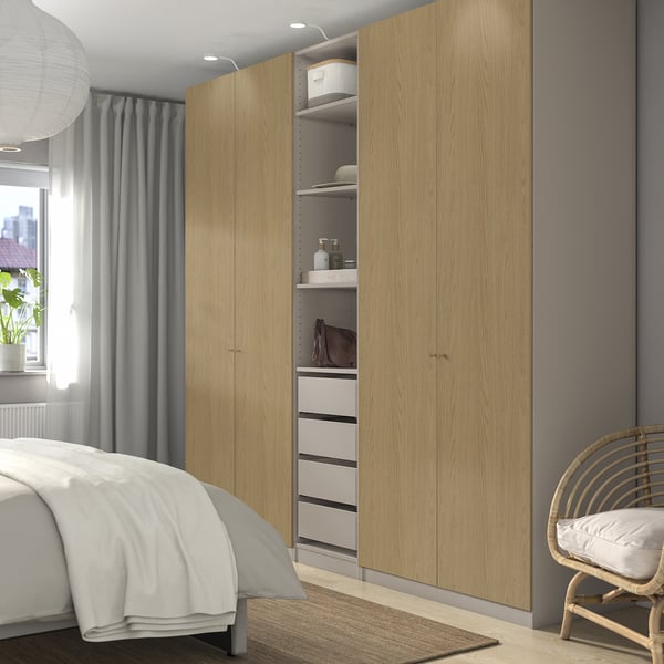 Spacious bedroom with light wood PAX wardrobe, open section, white rug, wicker chair, window, grey curtains, and bed with white linens.
