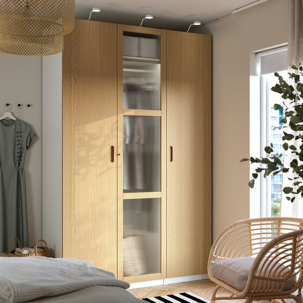 Modern bedroom featuring a large light brown PAX wardrobe with glass doors, next to a rattan chair and a bed with green blanket. Bright and spacious.