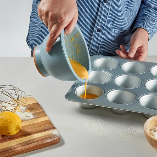 Person pouring yellow batter from a whisk bowl into a muffin tin.