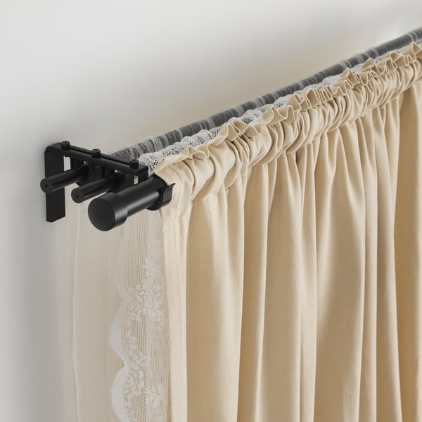 Black metal curtain rod with adjustable length, holding beige curtains with a lace trim, mounted on a wall.