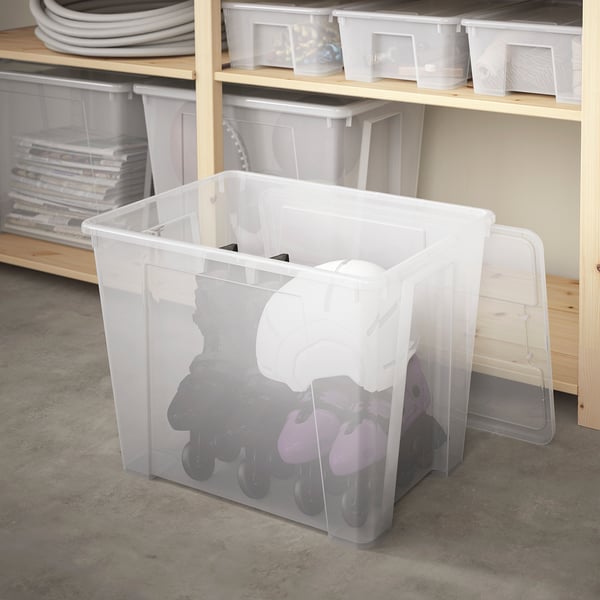 A clear plastic SAMLA box on the floor, storing rollerblades. Or SAMLA storage box on floor holding rollerblades.