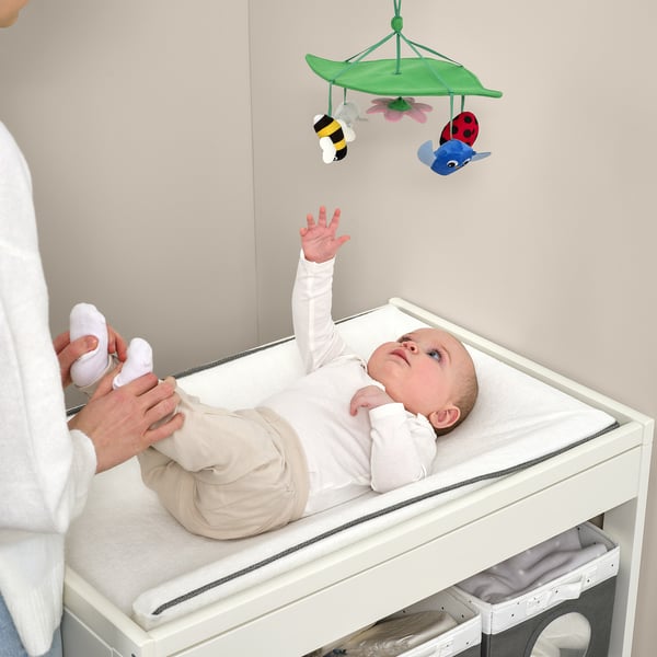 Baby reaching for hanging mobile with colourful bugs above white changing table. Adult helps babys foot.
