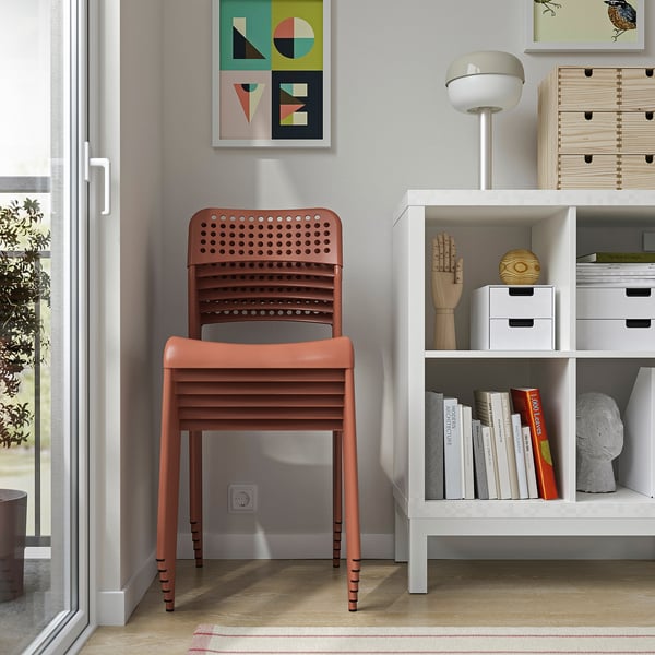Orange plastic chair stacked on another, next to a white bookshelf with various items, including books and decorative objects.