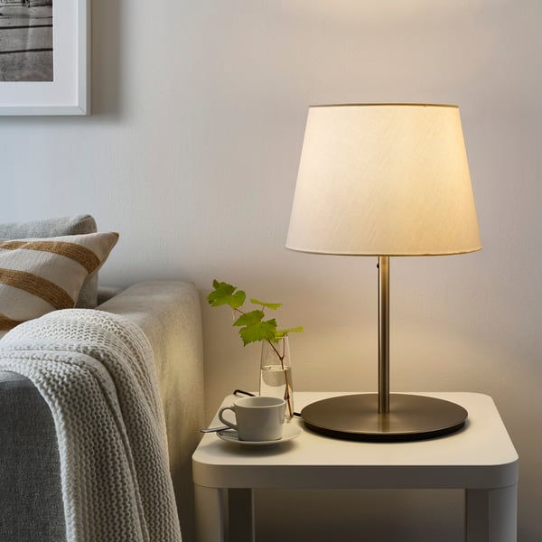 Minimalist living room with SKAFTET lamp, beige shade on side table. Next to a sofa draped with a white blanket and a small plant.