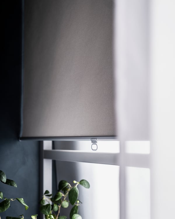 A grey roller blind, partially open, on a white window with a plant nearby.