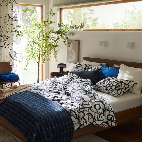 Bedroom with SKOGSKOVALL blue jacquard cotton bedspread, geometric pattern, large windows, lush plants, wooden furniture, cosy & inviting.