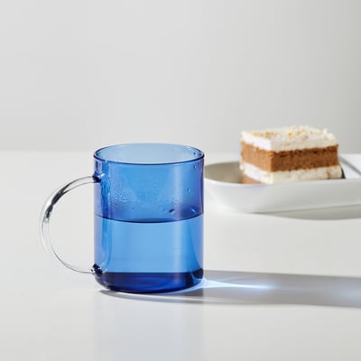 Blue glass mug with handle, half-filled with water, sits near a slice of cake on a white tray.