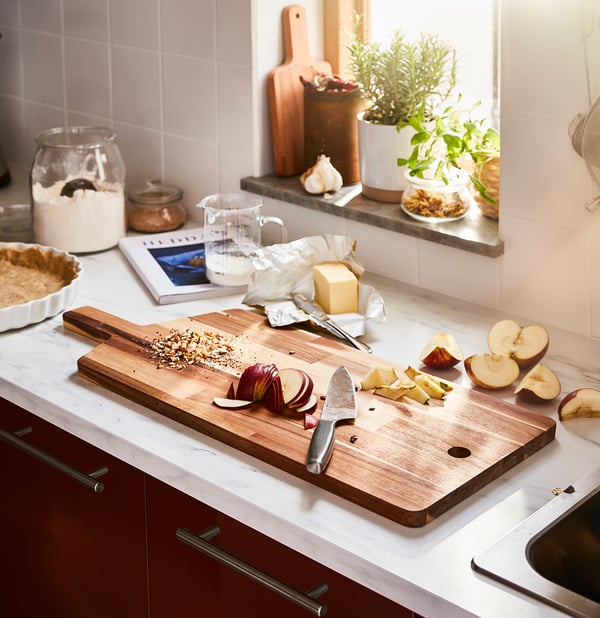A wooden cutting board with knife and apple slices sits on a white kitchen countertop, surrounded by baking ingredients.